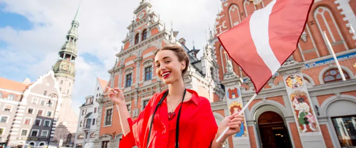  In Latvia, a woman dressed in red stands before a building, holding a flag that represents her country's heritage.